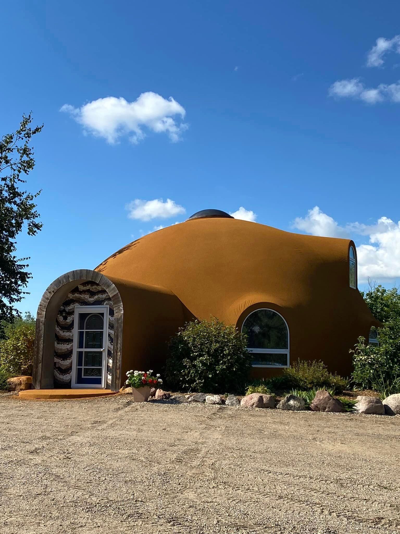 Beige dome-shaped house with arched entryway, small windows, gravel driveway, and blue sky.