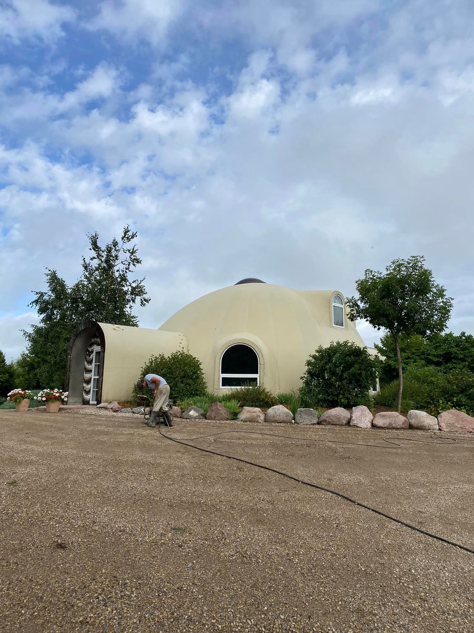 Beige geodesic dome house with surrounding landscaping, cloudy sky.