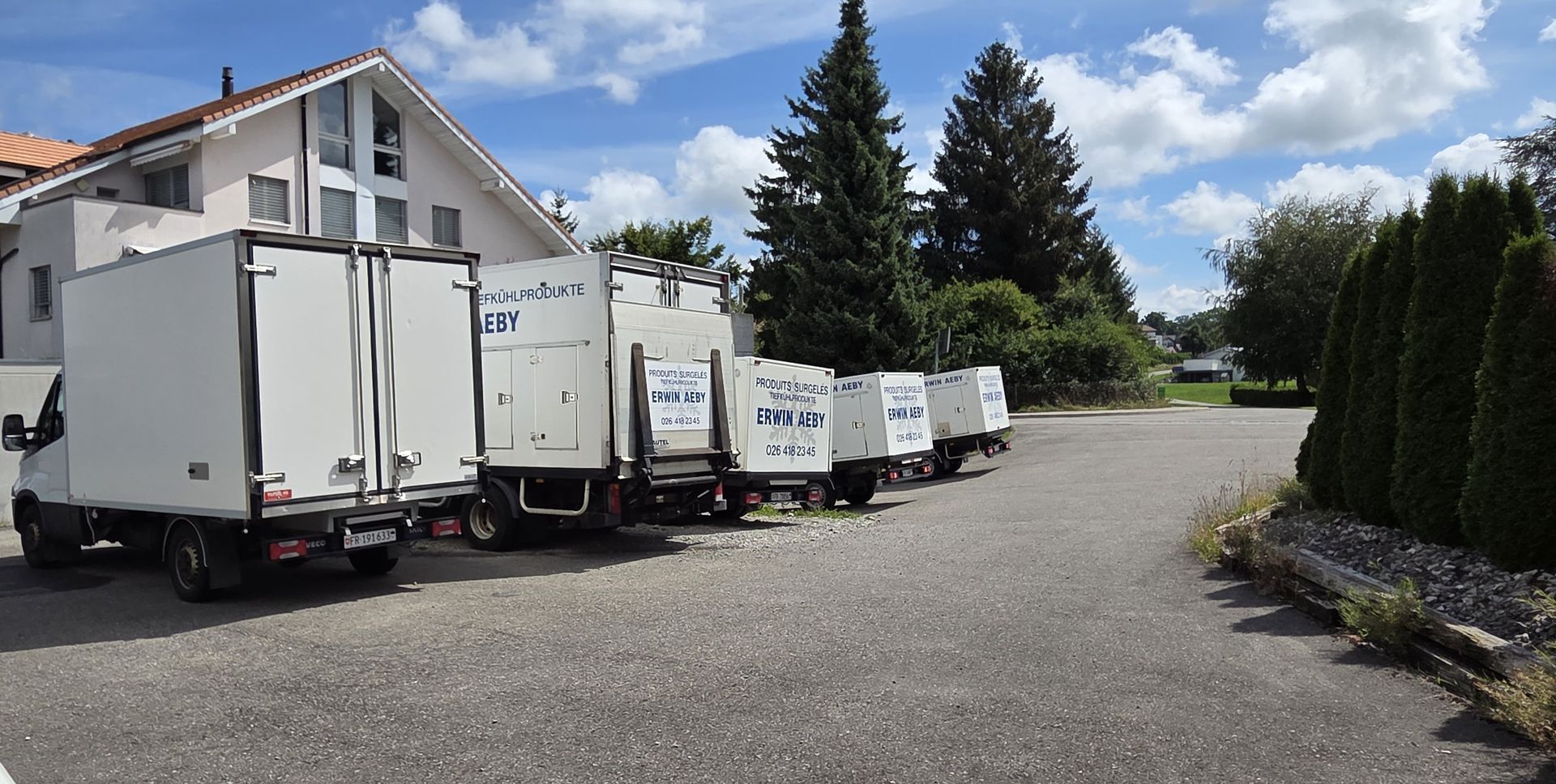 Camions de livraison blancs garés sur du gravier devant un bâtiment et des arbres par une journée ensoleillée.