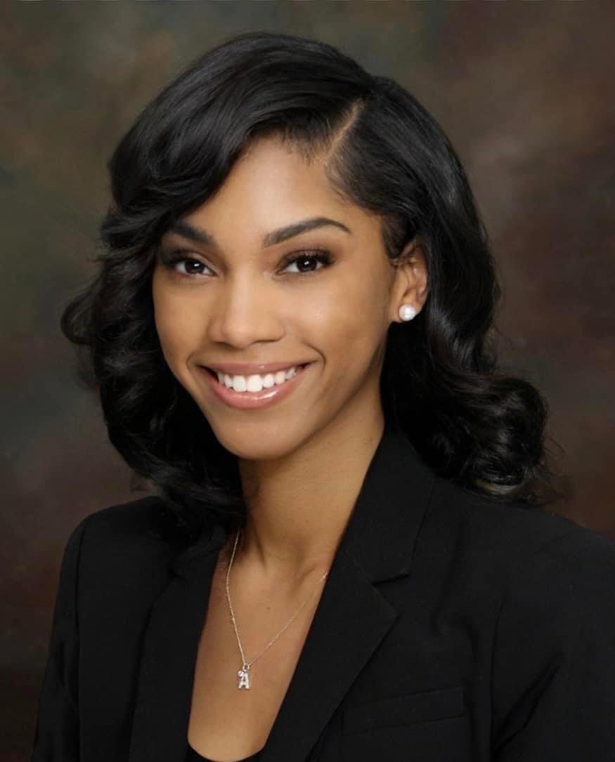A woman wearing a black jacket and a necklace smiles for the camera