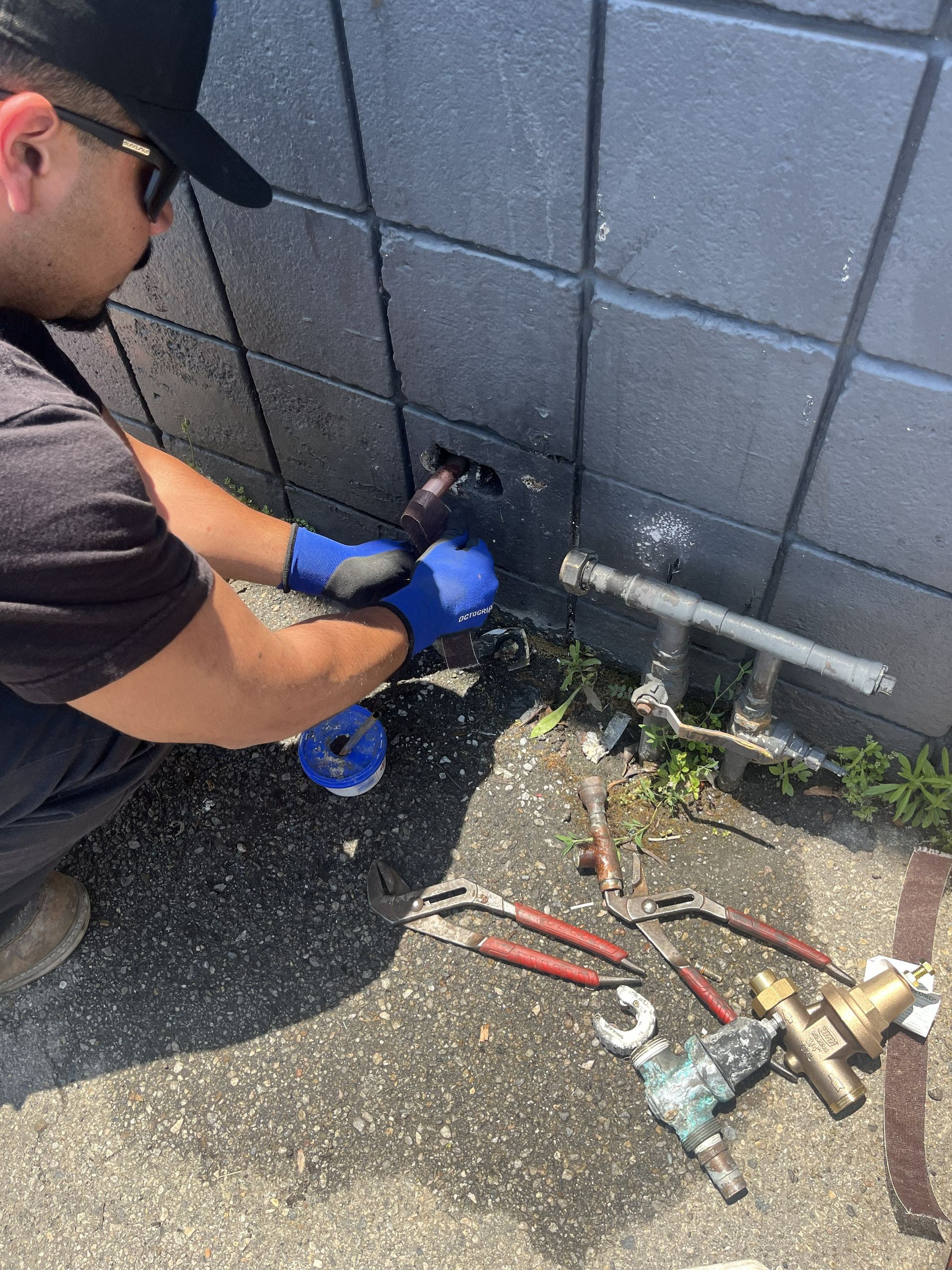 Worker using blue gloves repairs a pipe against a gray wall with tools on the ground.