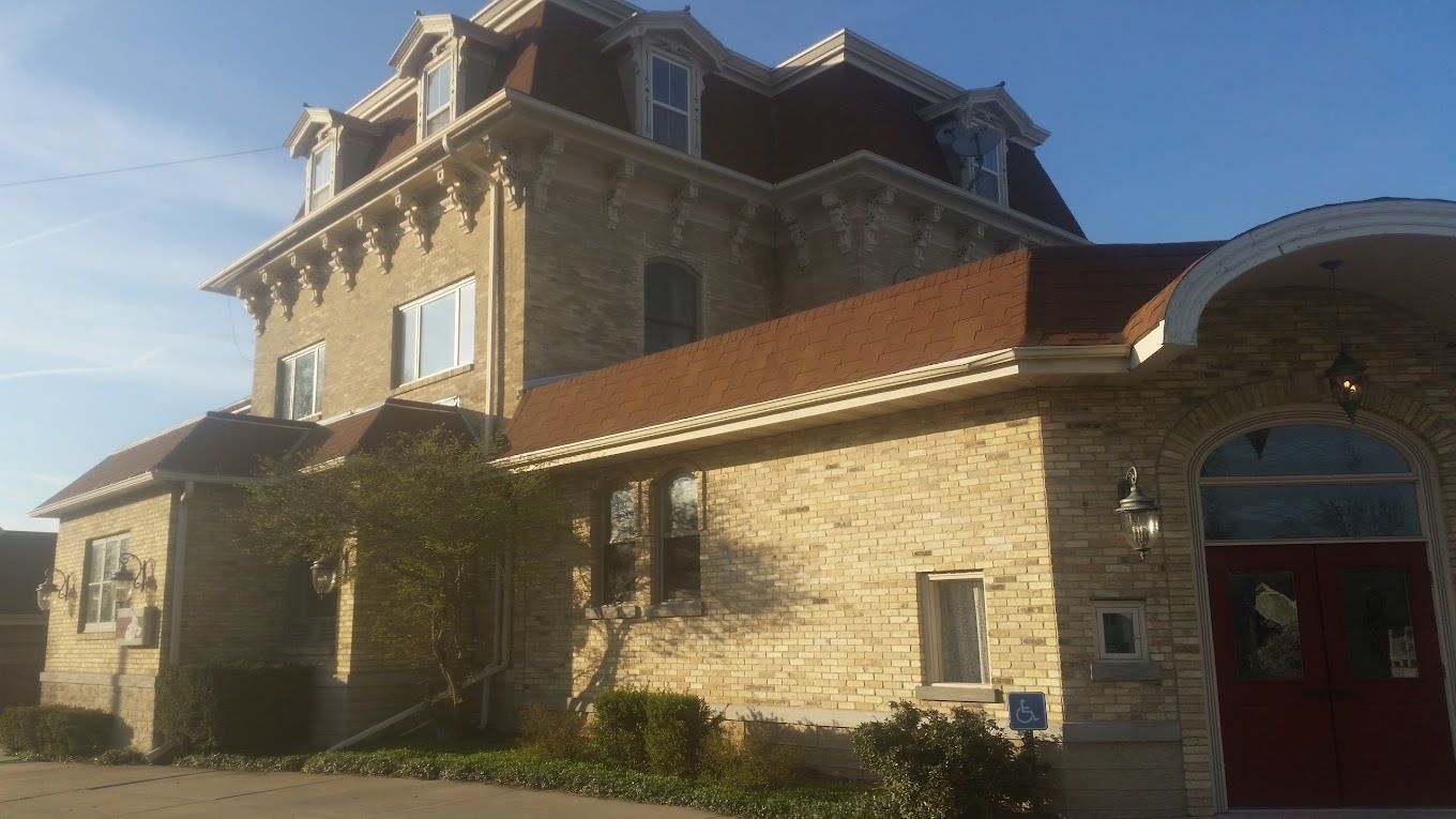 A beige stone building with a dark brown mansard roof and a red arched entrance door under a bright blue sky.