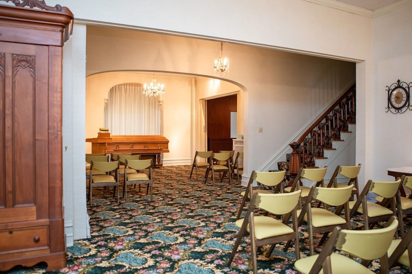 An interior view of a funeral home with rows of chairs facing an open casket on a platform, all set on floral carpet.