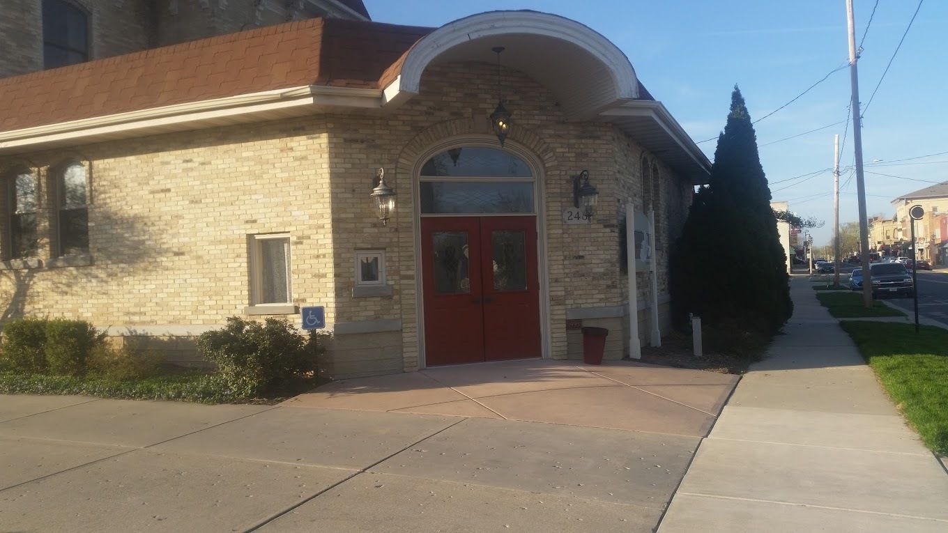 A stone building with a curved roof over a bright red double-door entrance next to a sidewalk on a sunny day.