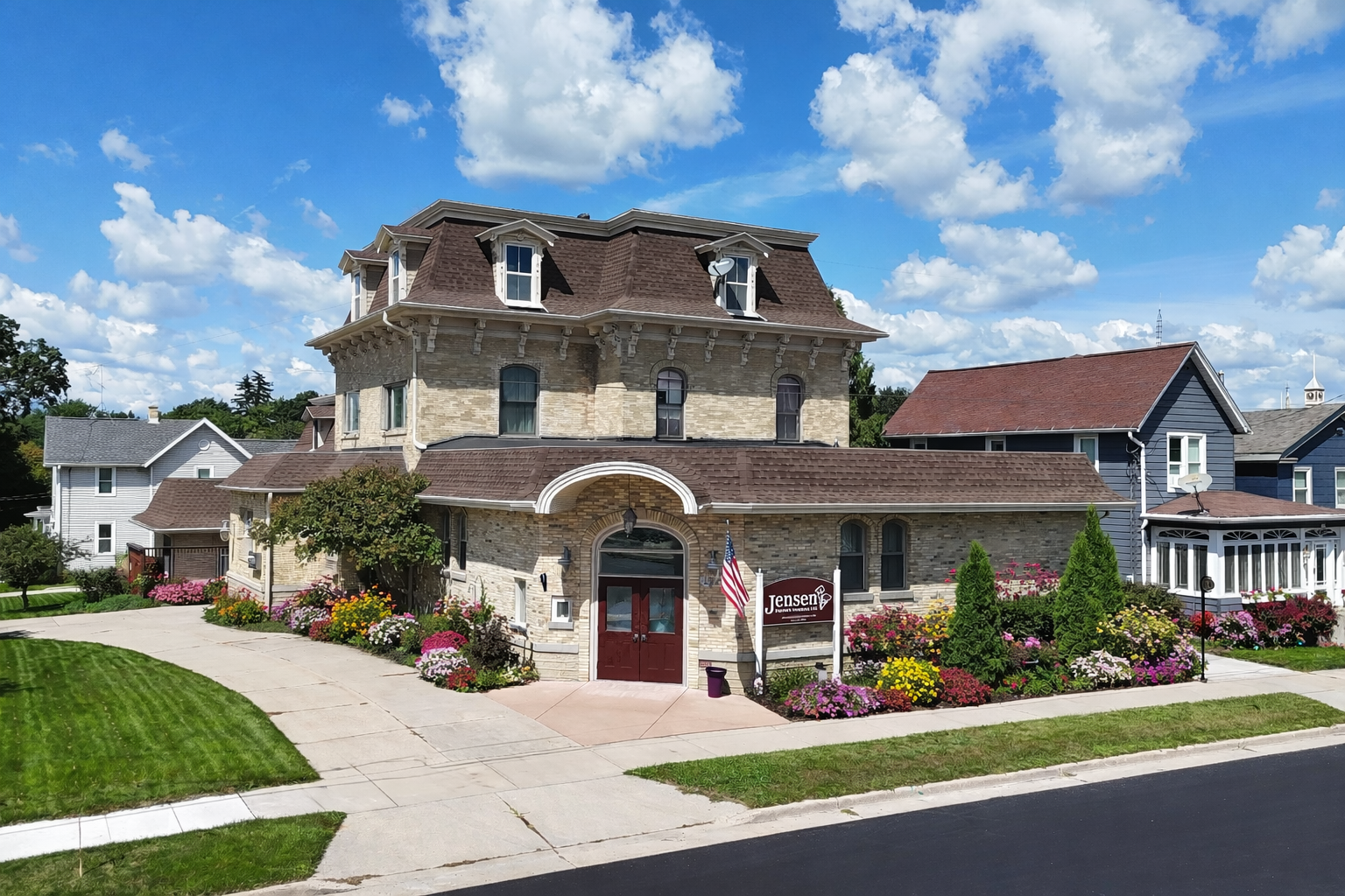 A multi-story stone building with a mansard roof, arched entryway, and landscaped garden under a bright, cloudy sky.