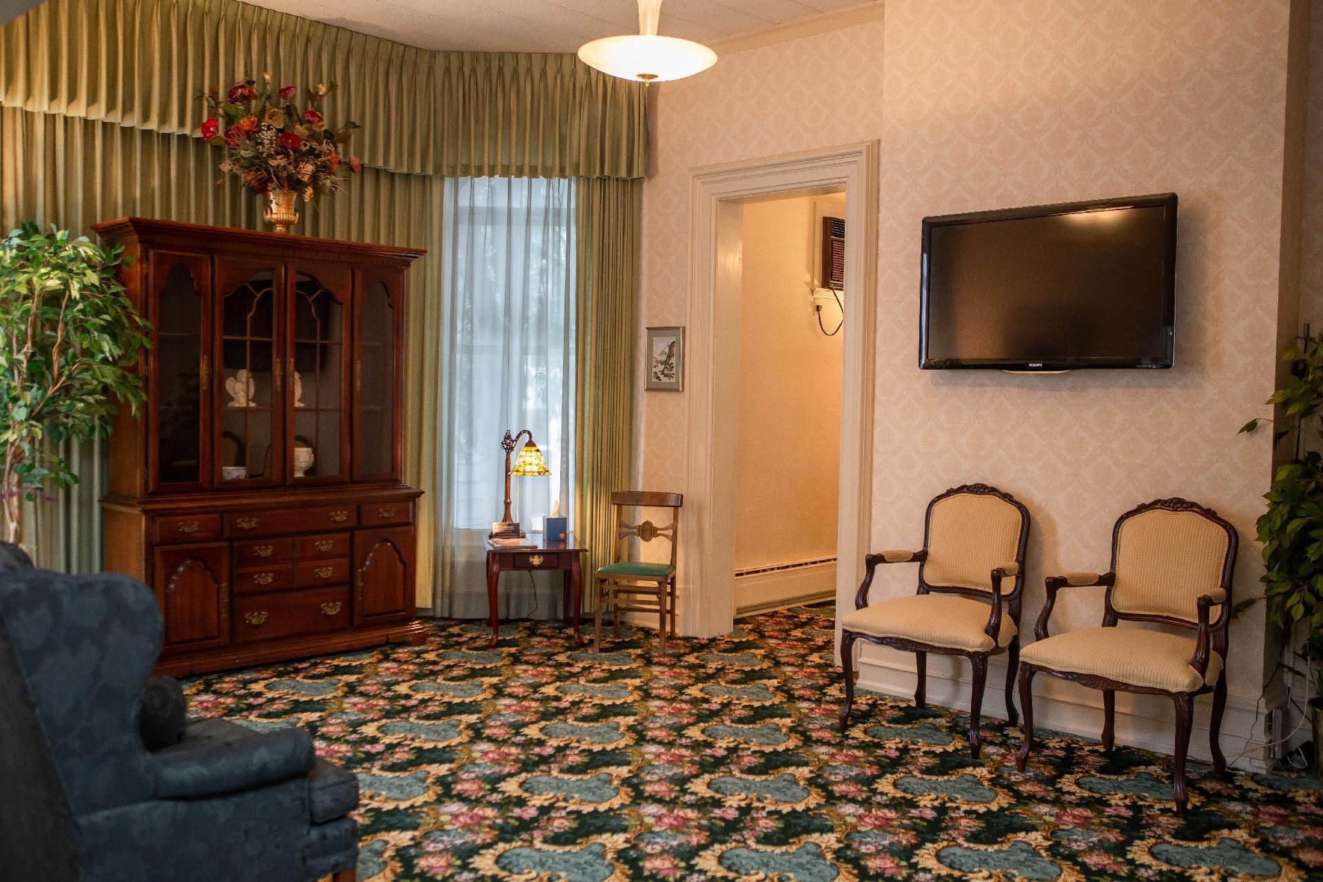 A formal room with a dark wood hutch, floral carpet, two upholstered chairs, and a wall-mounted television.