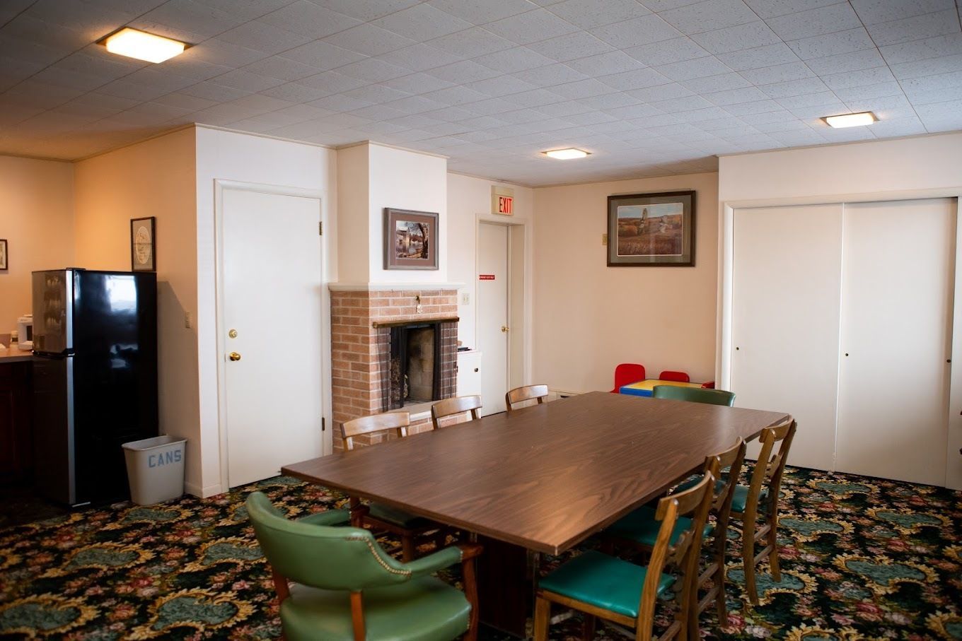 A meeting room with a large wooden table, green chairs, a brick fireplace, a black refrigerator, and floral carpeting.