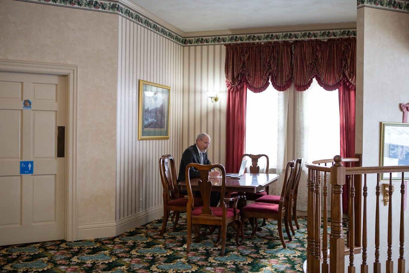 A person sits at a wooden table in a room with striped wallpaper and floral carpet, near a window with red curtains.