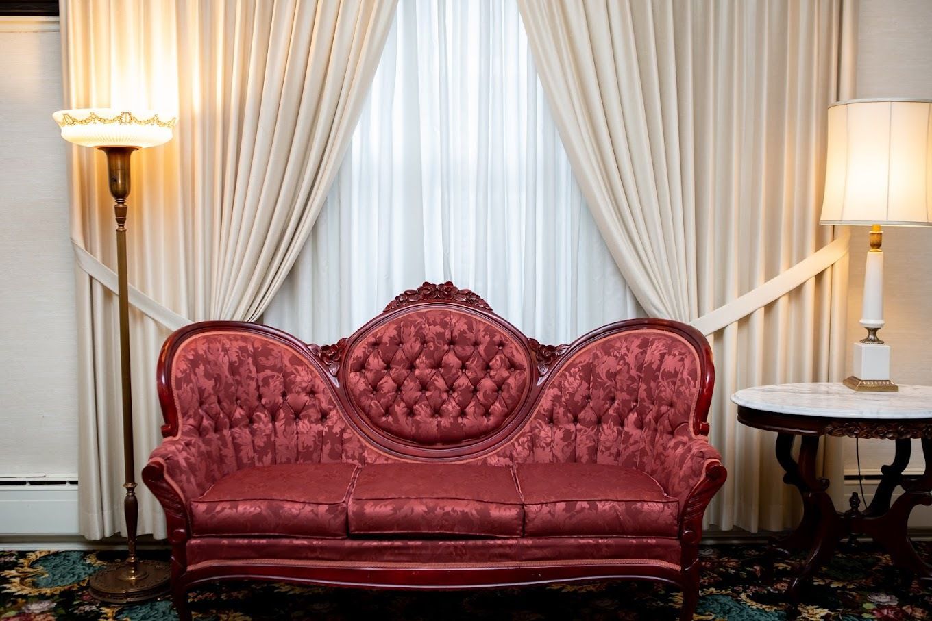 A red tufted Victorian-style sofa sits between two lamps against curtains in a room with a patterned floor.