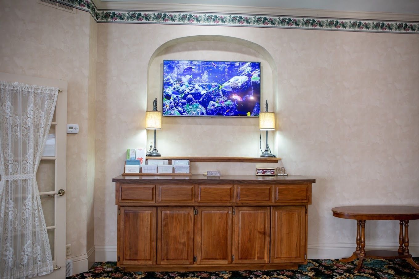 A wooden sideboard stands below a television in an arched wall niche, with matching lamps and a table to the right.