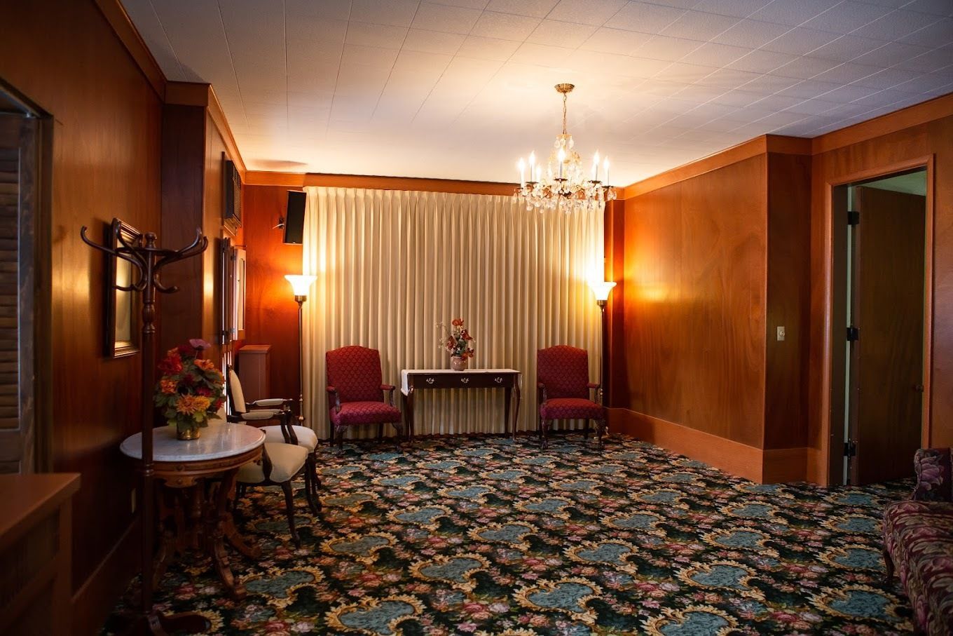 A room with patterned carpet, wood-paneled walls, two red chairs flanking a table, a chandelier, and a side table.