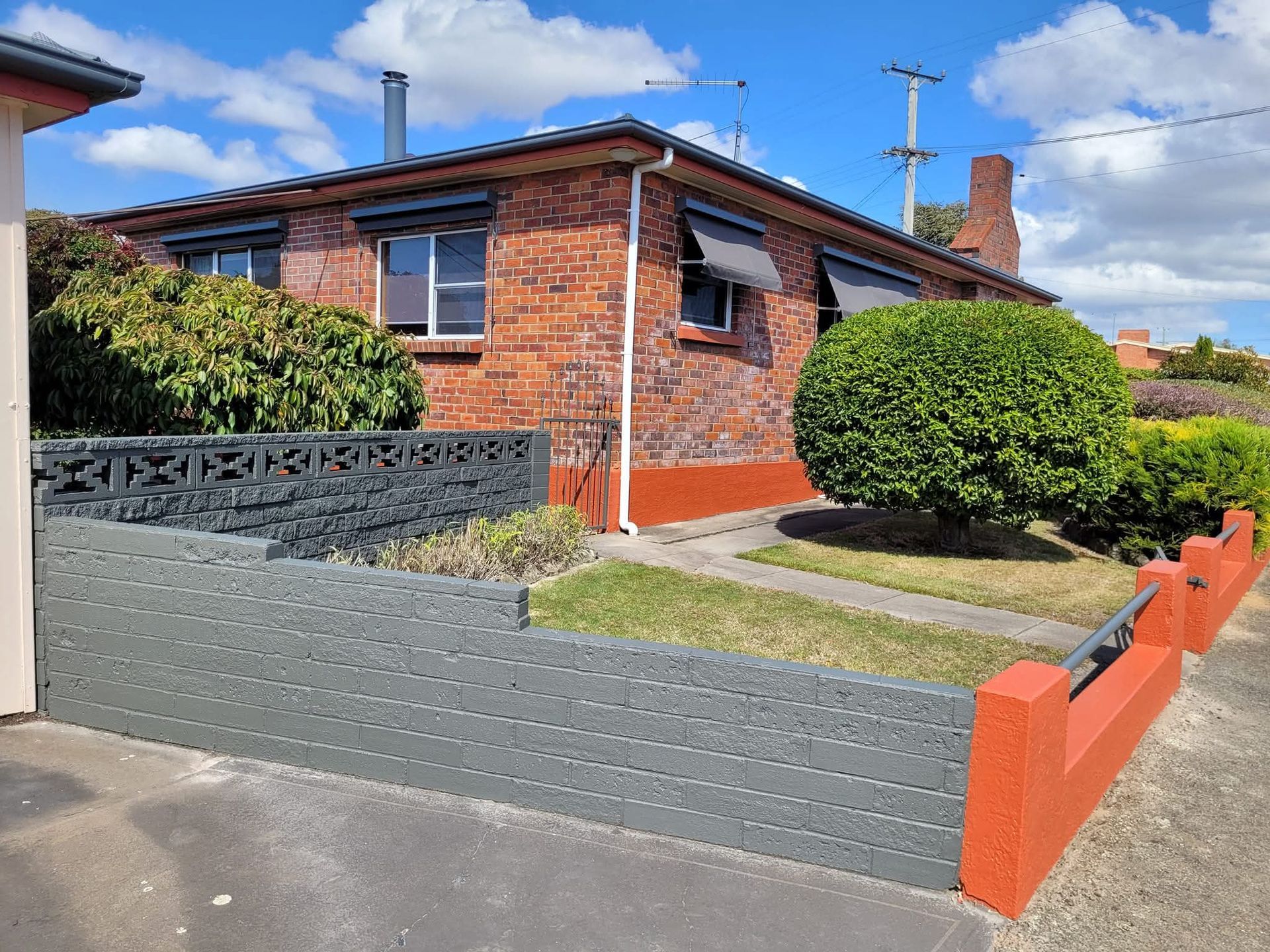 Brick bungalow with red trim, front garden, and driveway behind a low gray and orange wall under a blue sky