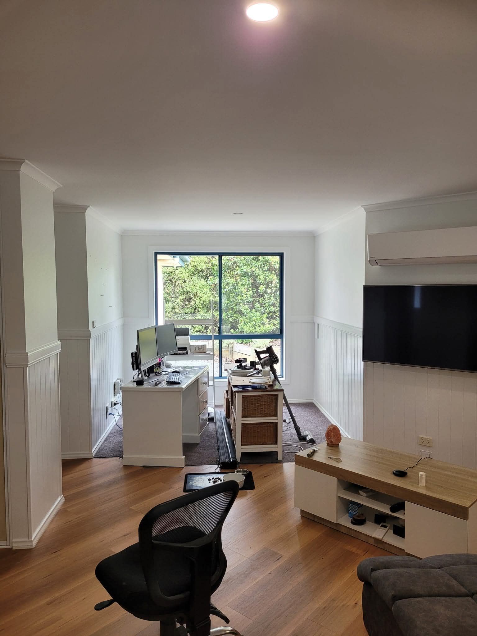 Bright living room with desk by window, hardwood floors, black chair, and TV on a white wall