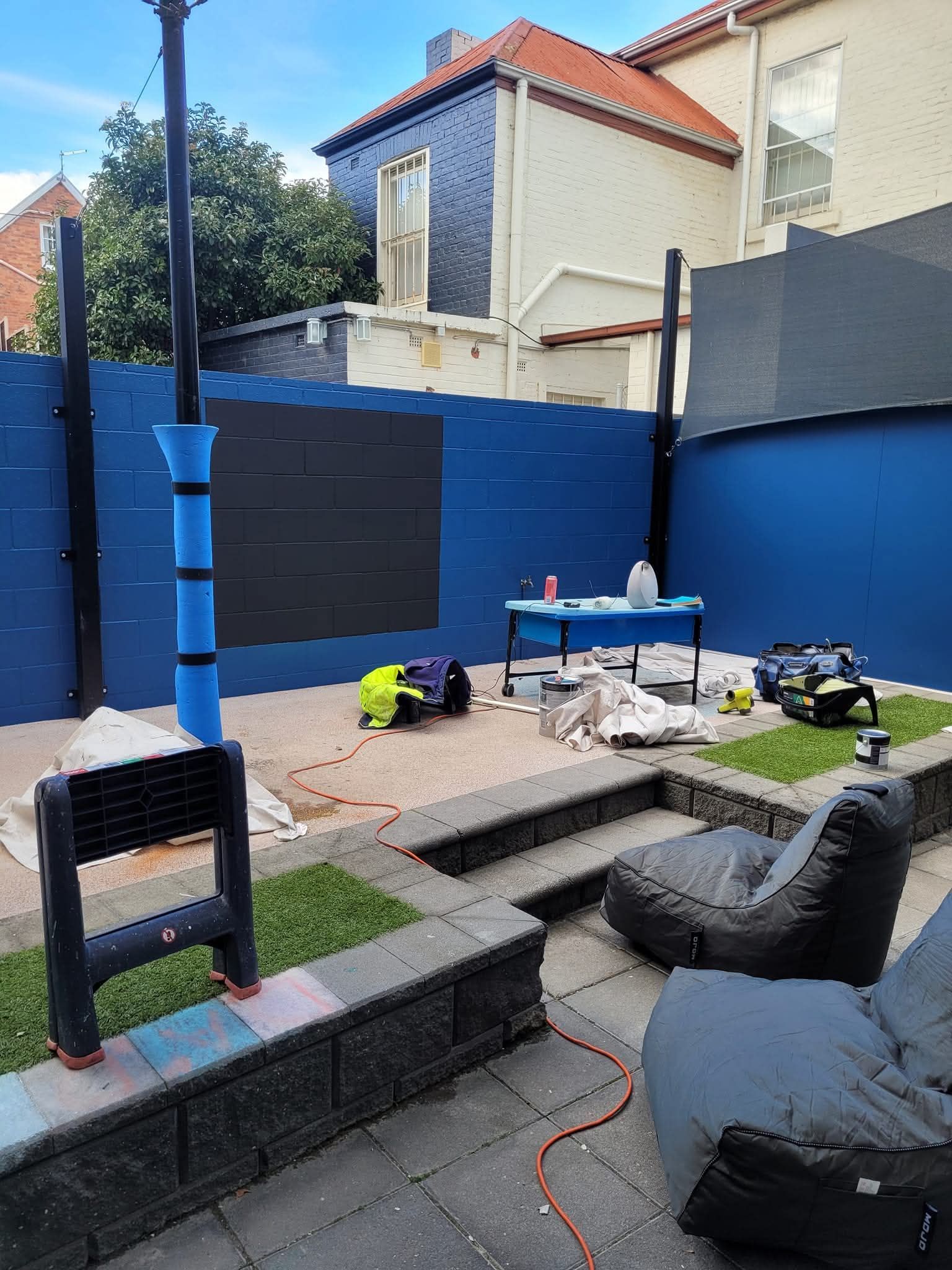 Small backyard patio with blue walls, gray seating, a fire pit, and tools scattered on the ground.