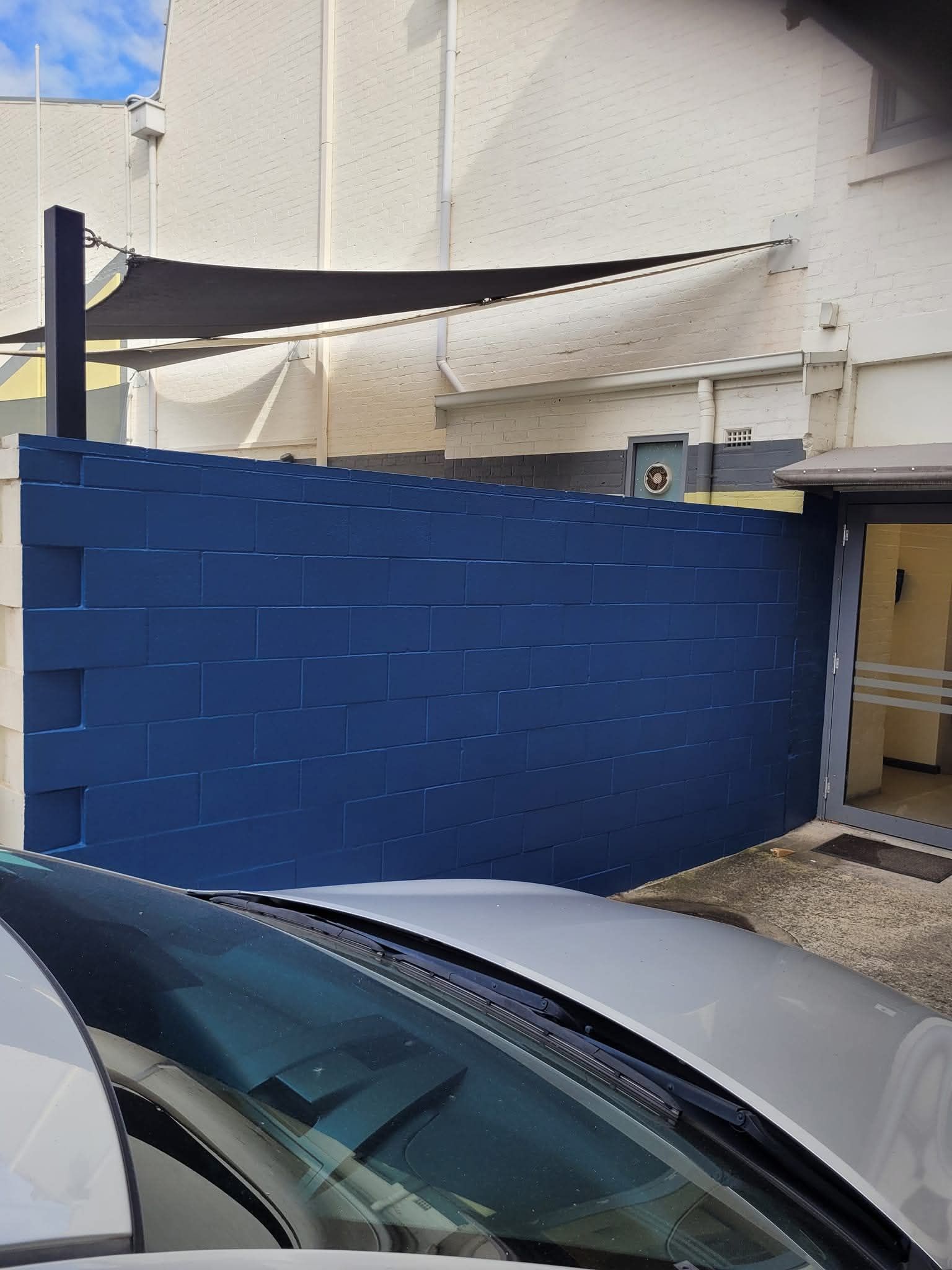 Blue brick wall beside a white building, with parked cars in the foreground and a small gate on the right.