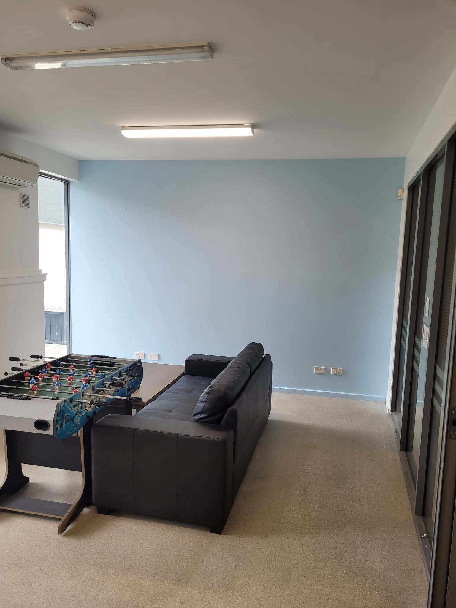 Empty living room with beige carpet, blue wall, black sofa, and a small table by the window.