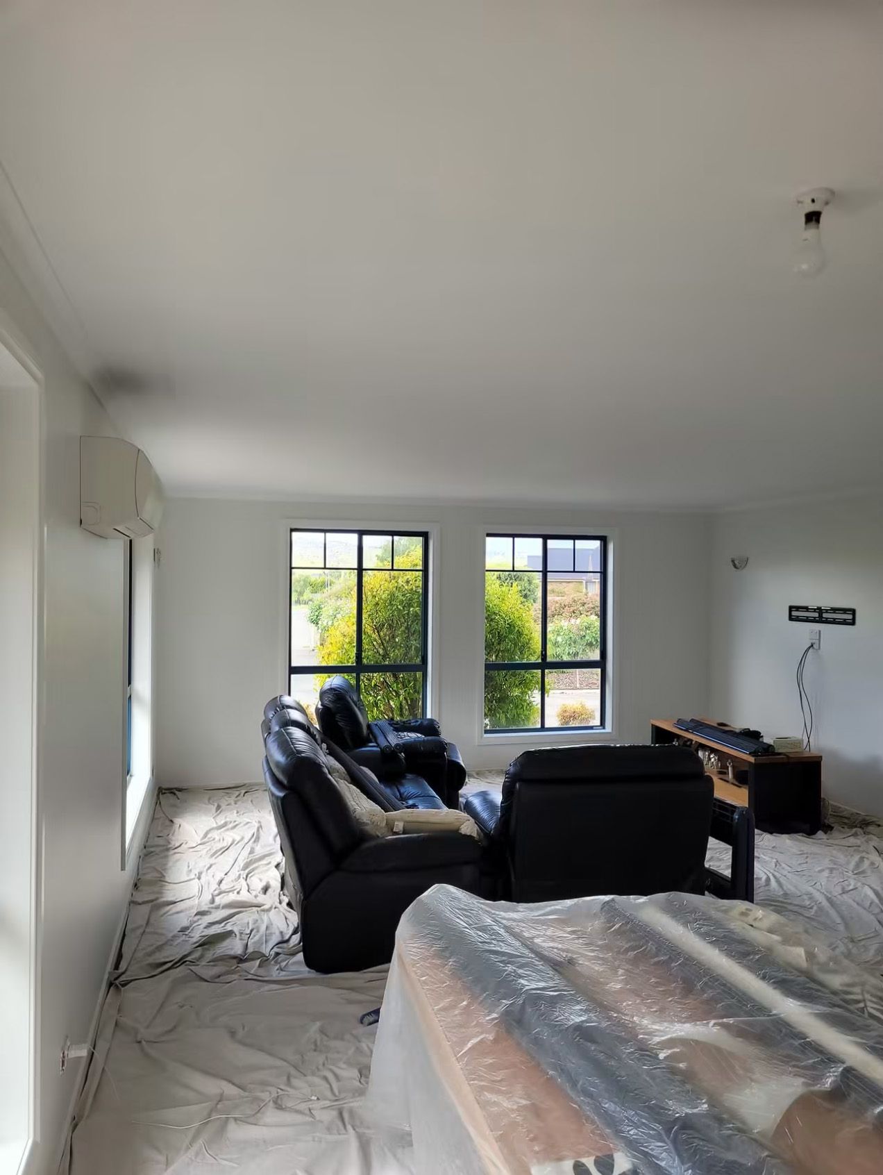 Bright living room with two sofas, large windows, and furniture wrapped in plastic in the foreground.