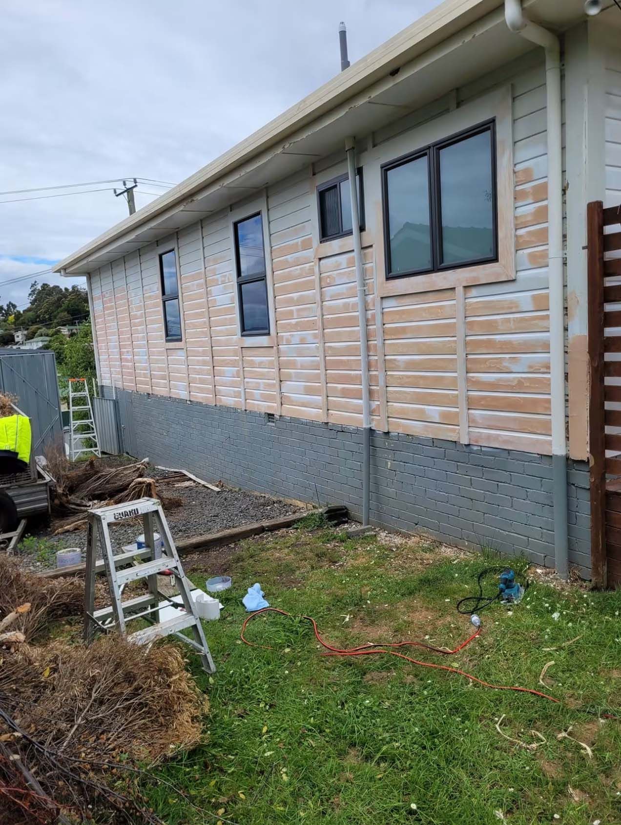 Side of a house under renovation with unfinished siding, windows, ladder, and scattered tools in the yard.