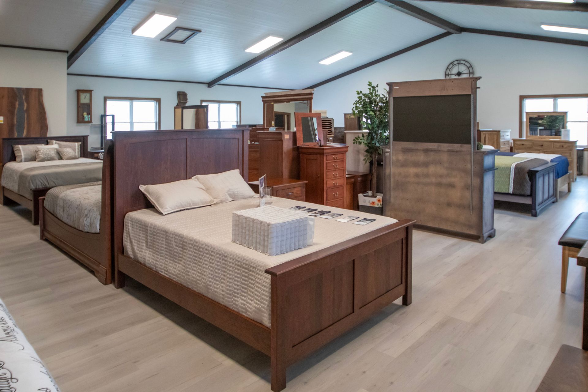 Bedroom furniture showroom with wooden beds, dressers, and flooring. Natural light and high ceilings.