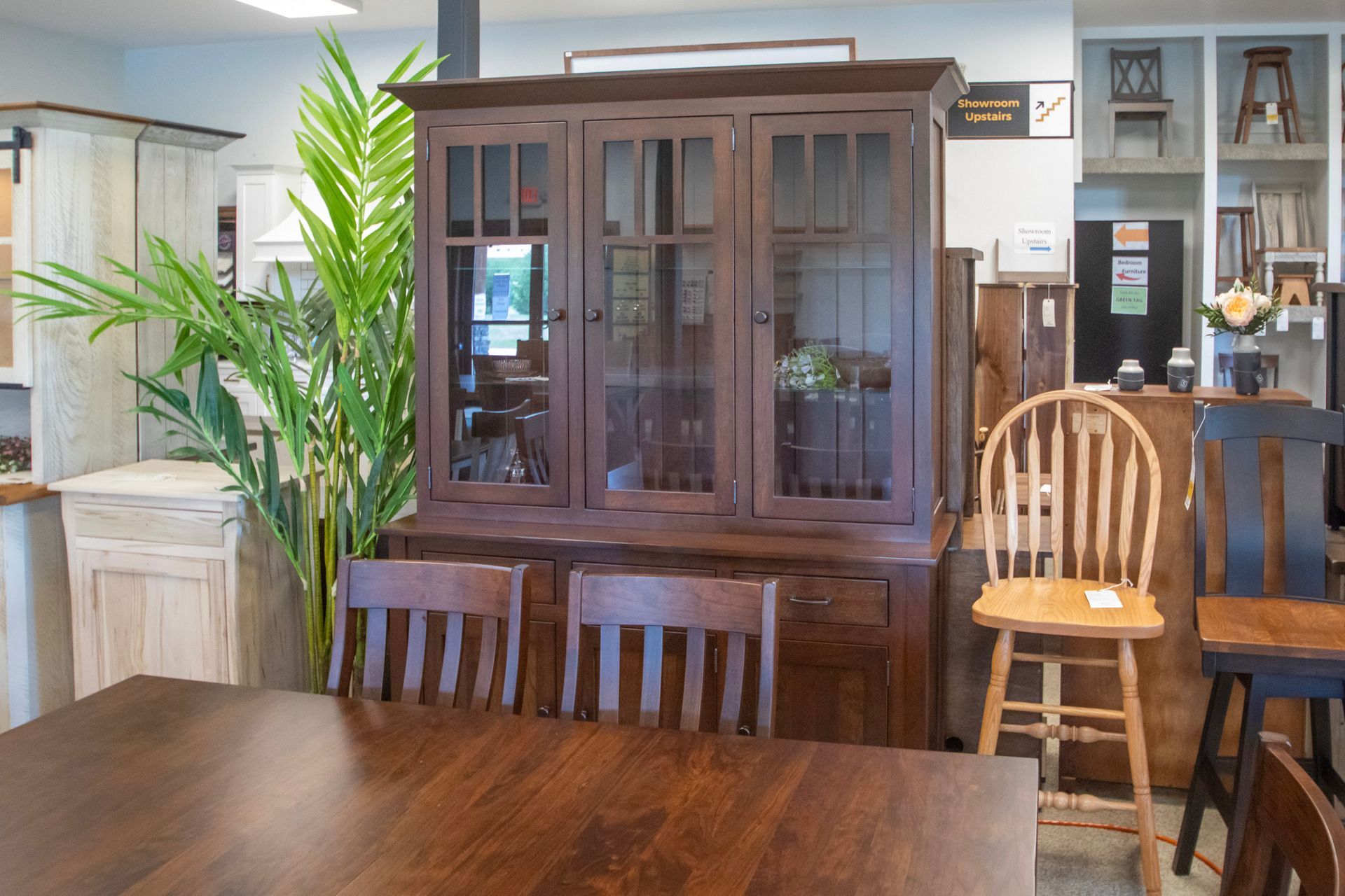 Dining room furniture display: wooden hutch with glass doors, table, chairs, and potted plant.