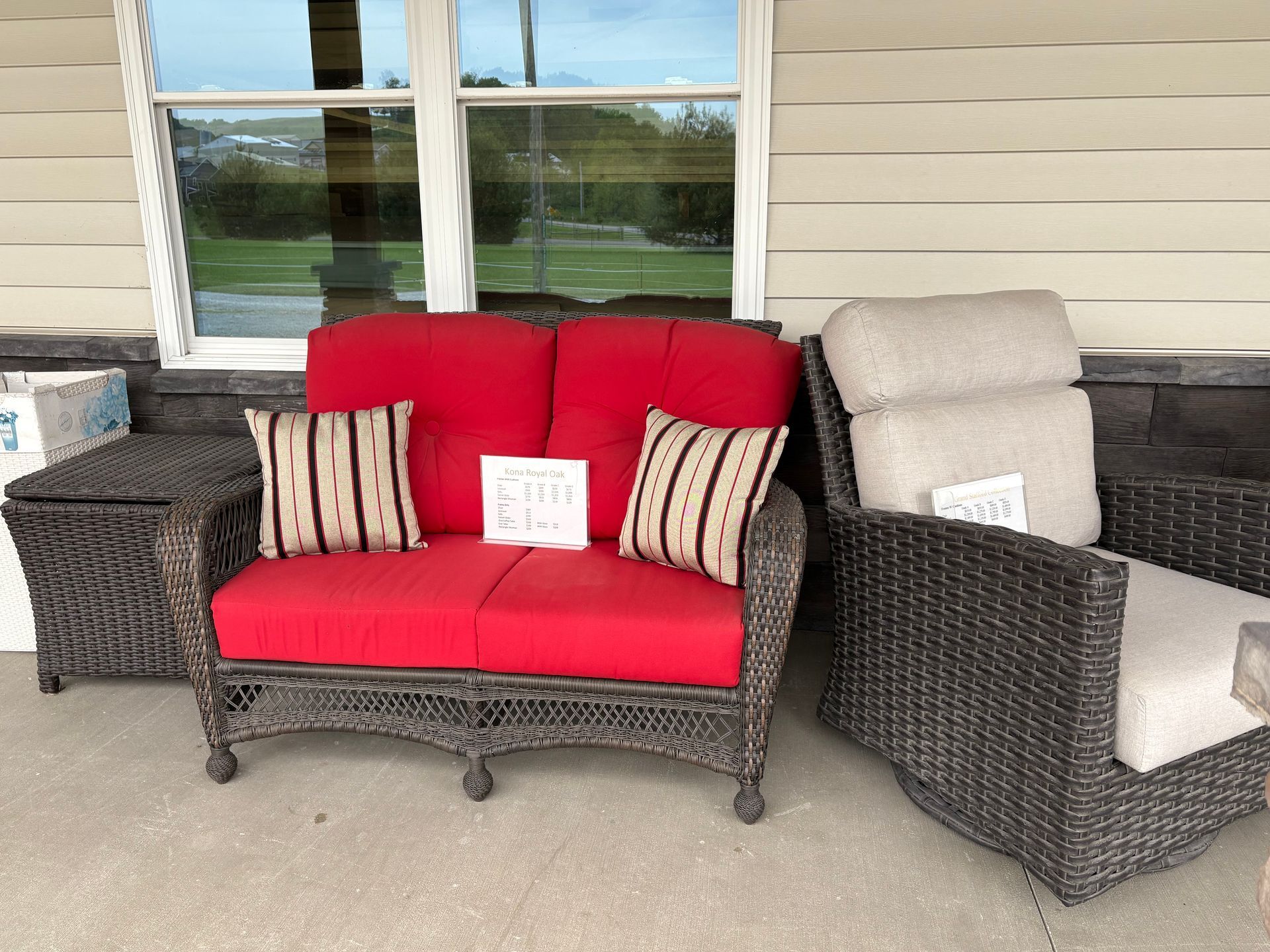 Wicker patio furniture: red loveseat with patterned pillows, beige chair, and small storage table sit on a porch.