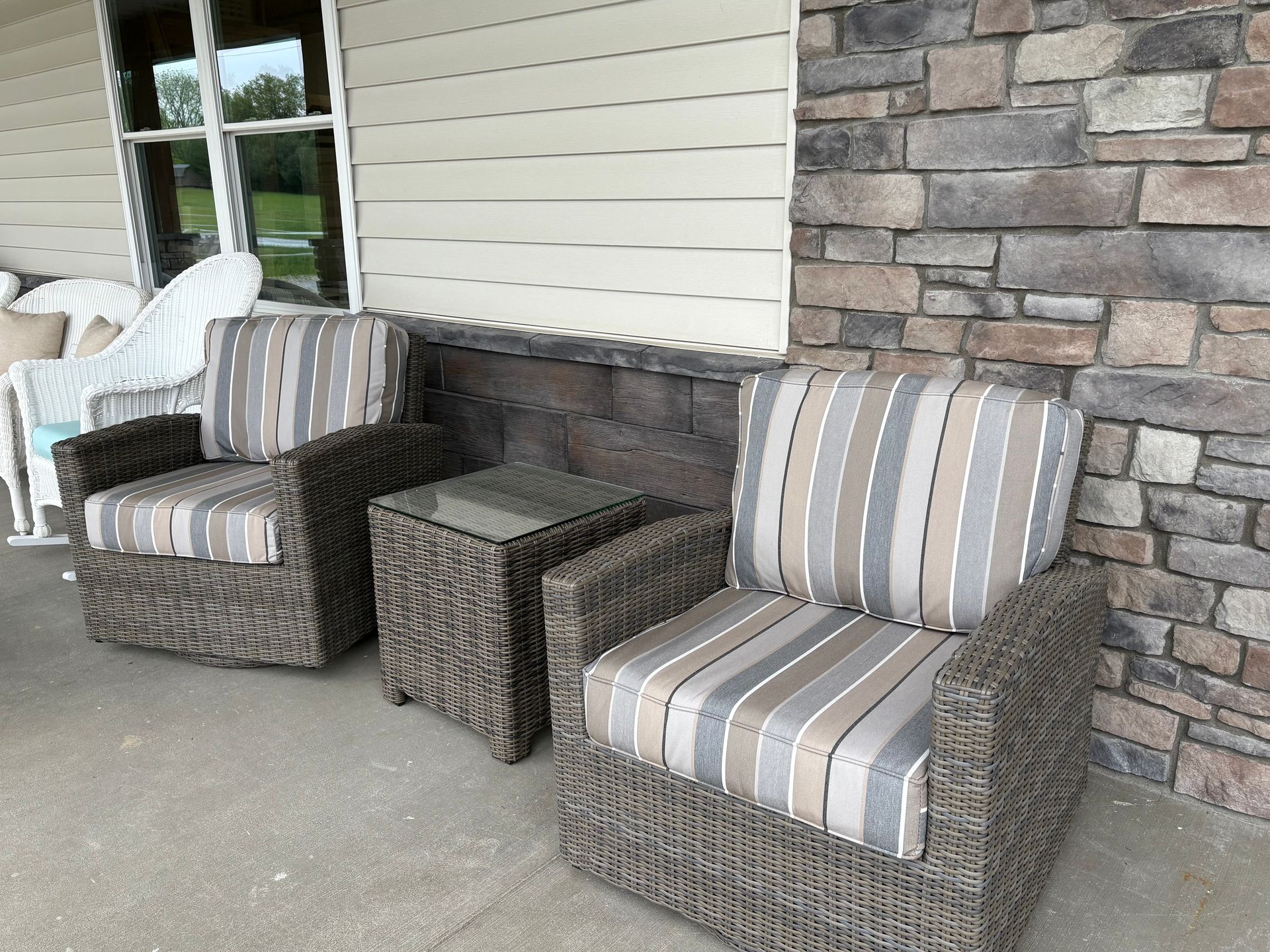 Two wicker chairs with striped cushions and a side table on a porch with stone accents.