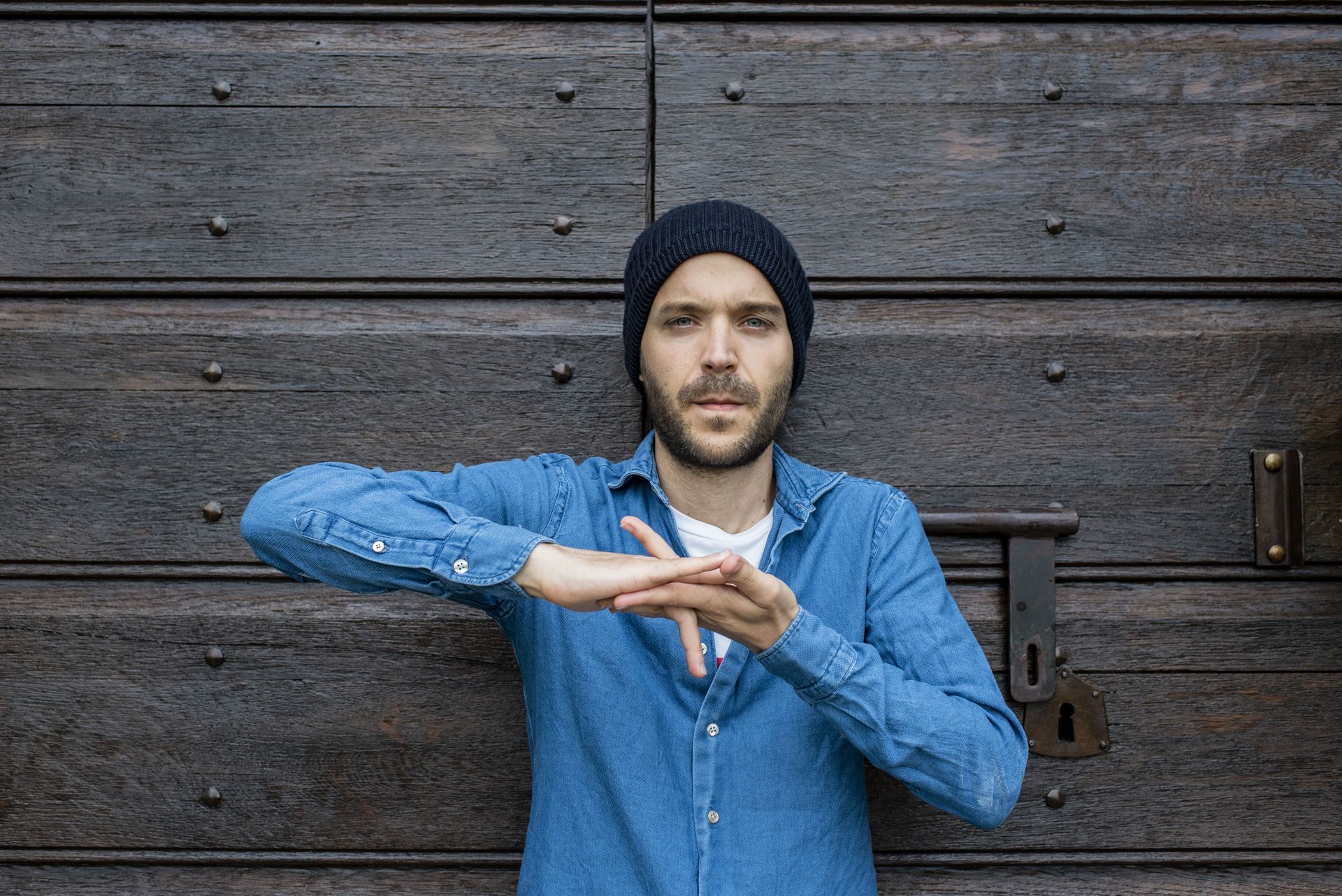 Una persona con una camicia blu e un berretto scuro è in piedi davanti a una porta di legno rustica, con le mani giunte davanti al petto.