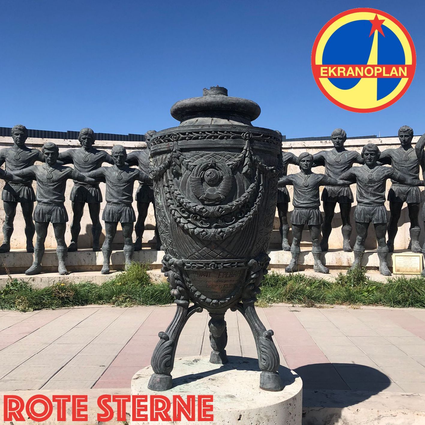 Un'urna metallica si erge davanti a una fila di statue di bronzo identiche, sullo sfondo di un cielo azzurro, con il logo 