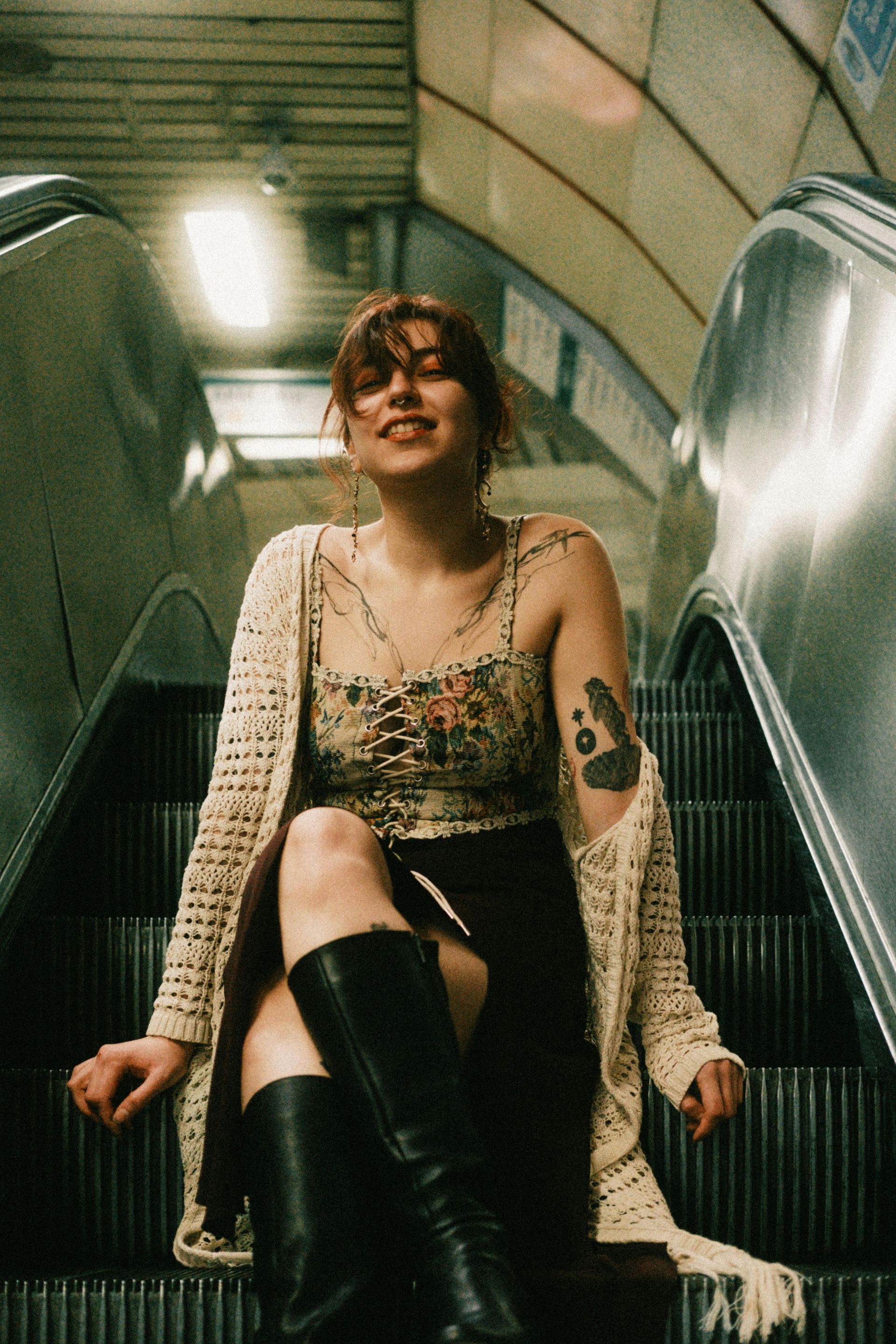 Una persona con un tatuaggio sul braccio è seduta su una scala mobile, indossa una maglietta a fiori, un cardigan lungo e stivali neri.