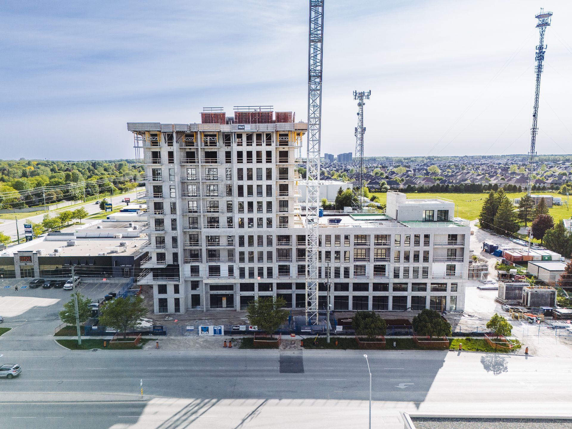 Multistory building under construction with cranes, seen from a street with trees and parking lot nearby