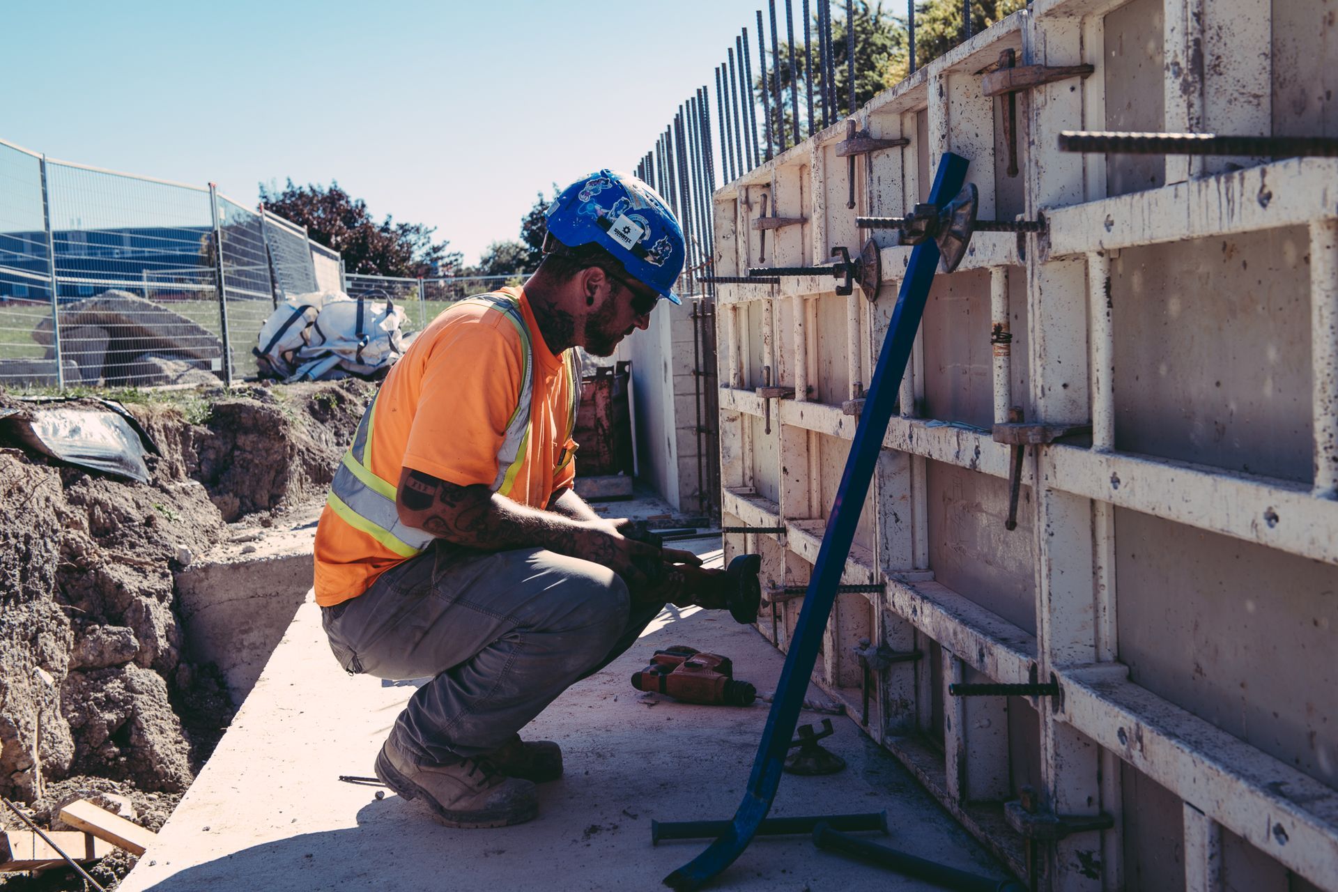 Construction worker in orange vest and blue hard hat kneeling beside a concrete wall, using a tool on site.