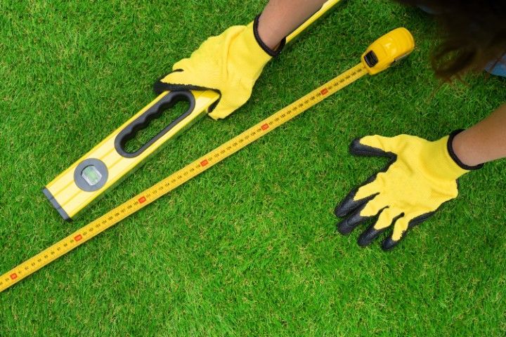 A person wearing yellow gardening gloves uses a spirit level and a measuring tape on green grass.