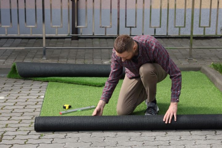 A person in a patterned shirt kneeling outdoors on a paved surface, unrolling a strip of artificial grass.