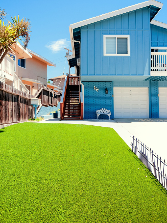 A bright blue, two-story house with a white garage sits behind a vibrant green lawn under a clear blue sky.