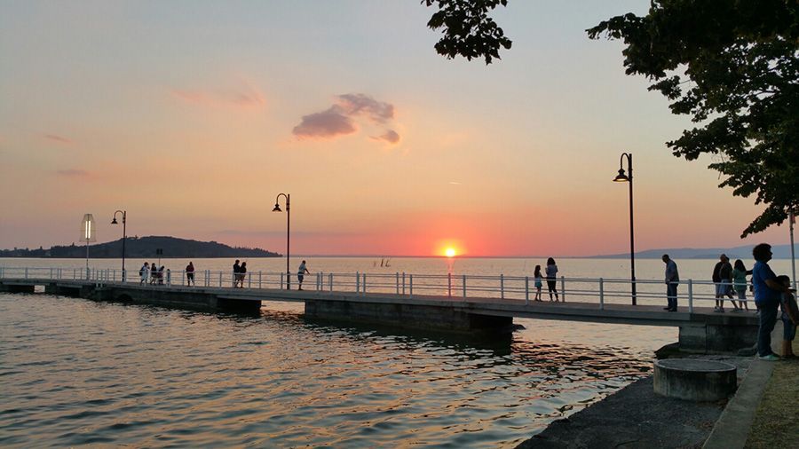 Tramonto su un molo sul lungomare, con gente che passeggia, acqua calma e lampioni che si stagliano contro un cielo rosa.