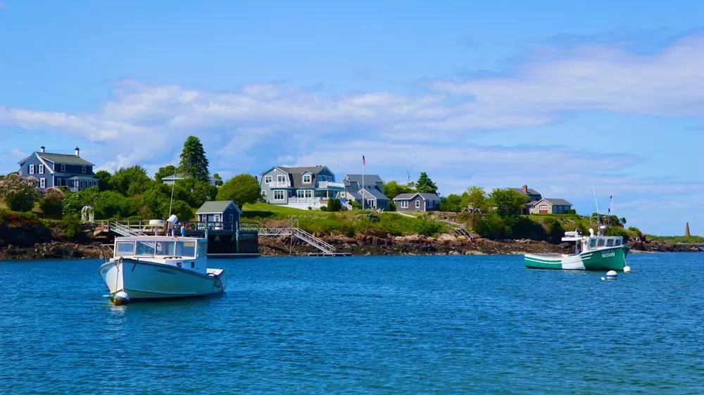 Harbor with two boats on blue water, rocky shoreline, and white houses under a partly cloudy sky
