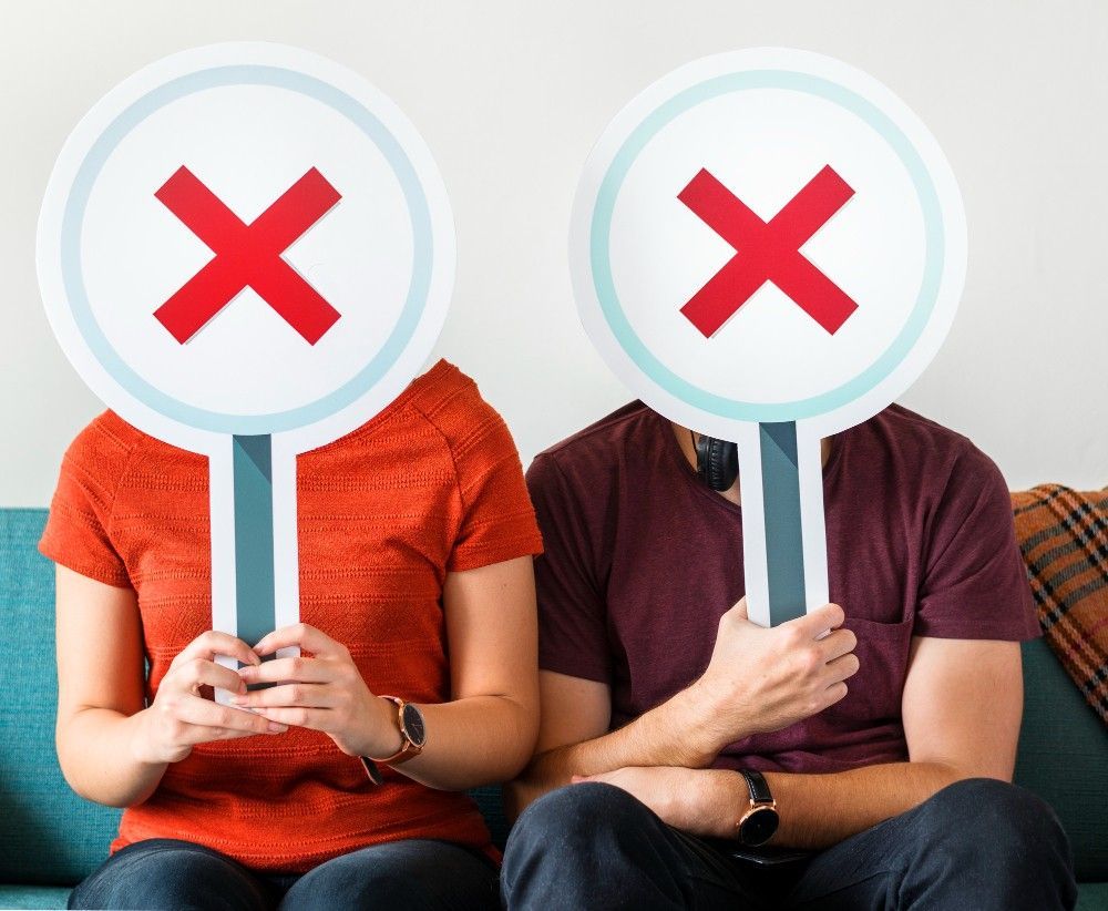 A man and a woman are sitting on a couch holding x signs in front of their faces.