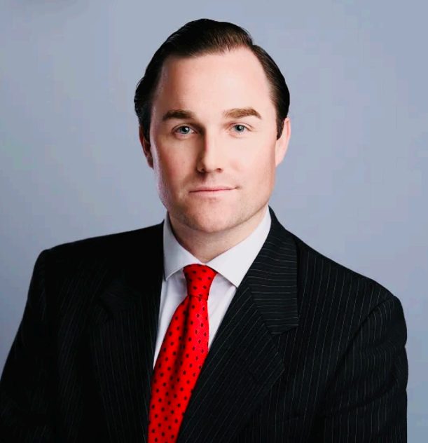 Professional headshot of a person in a black suit, white shirt, and red tie on a gray background