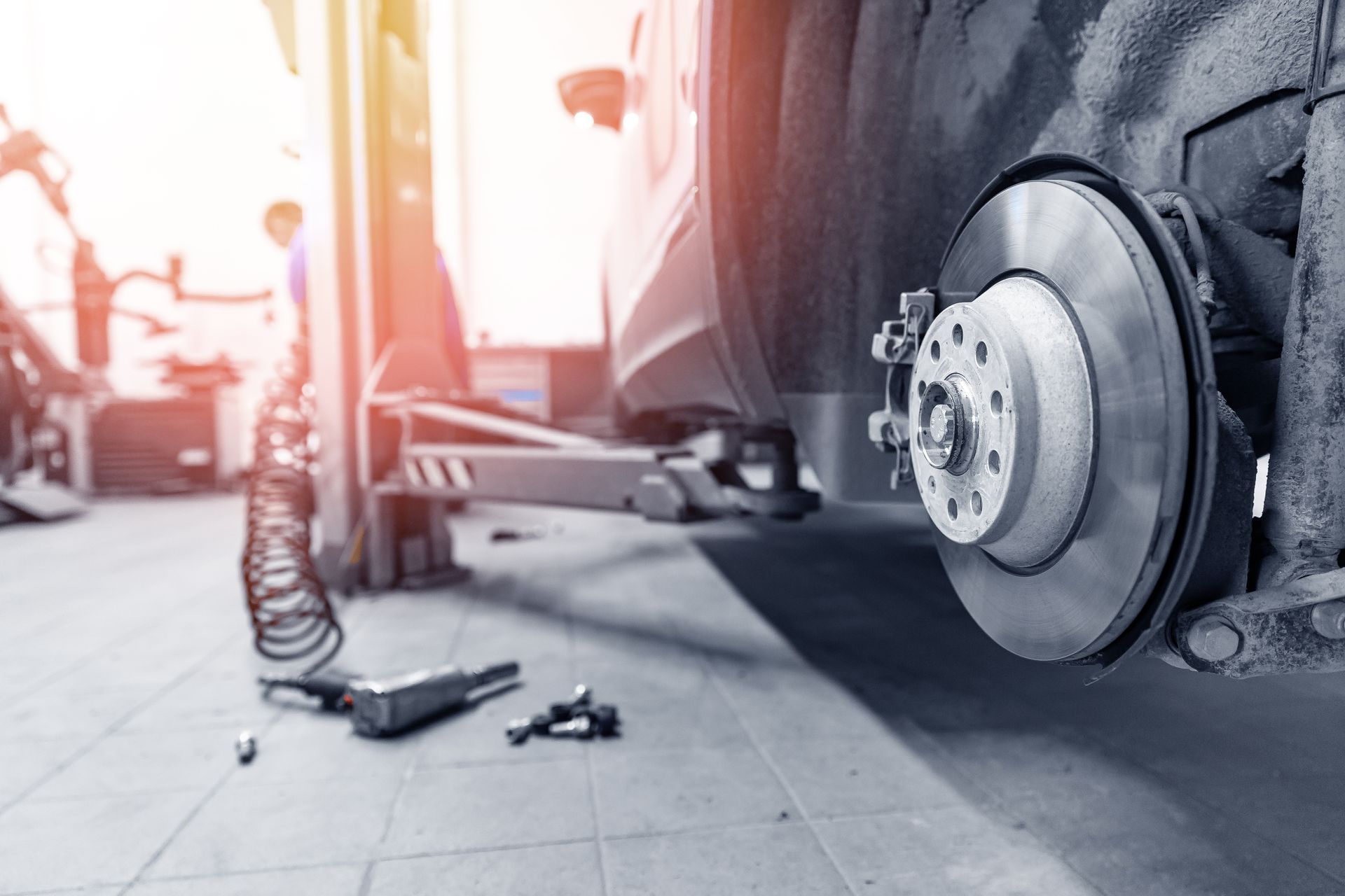 Car on jack with exposed brake rotor in a repair shop, tools on the floor.