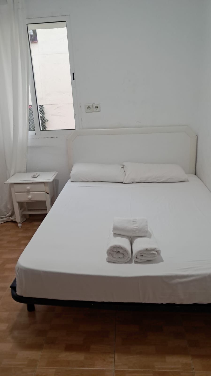 Dormitorio blanco con cama, mesita de noche, toallas y ventana.