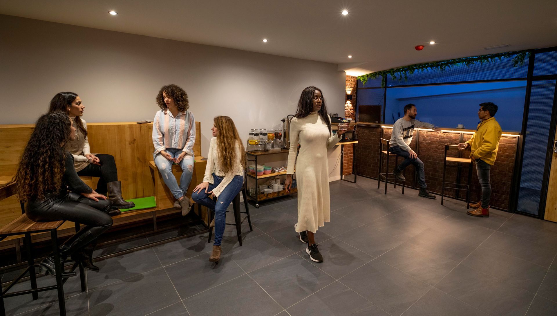 Gente socializando en un bar: cinco mujeres sentadas, una de pie y dos hombres en la barra. Iluminación tenue.
