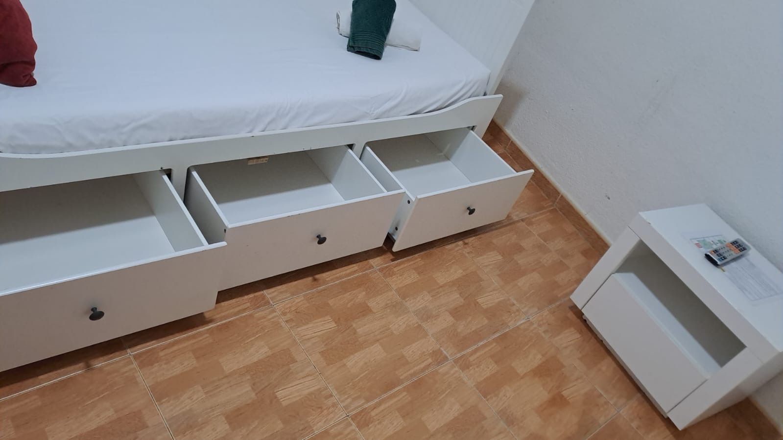 Cama blanca con cajones abiertos, al lado de una mesita de noche blanca sobre un piso de baldosas marrones.