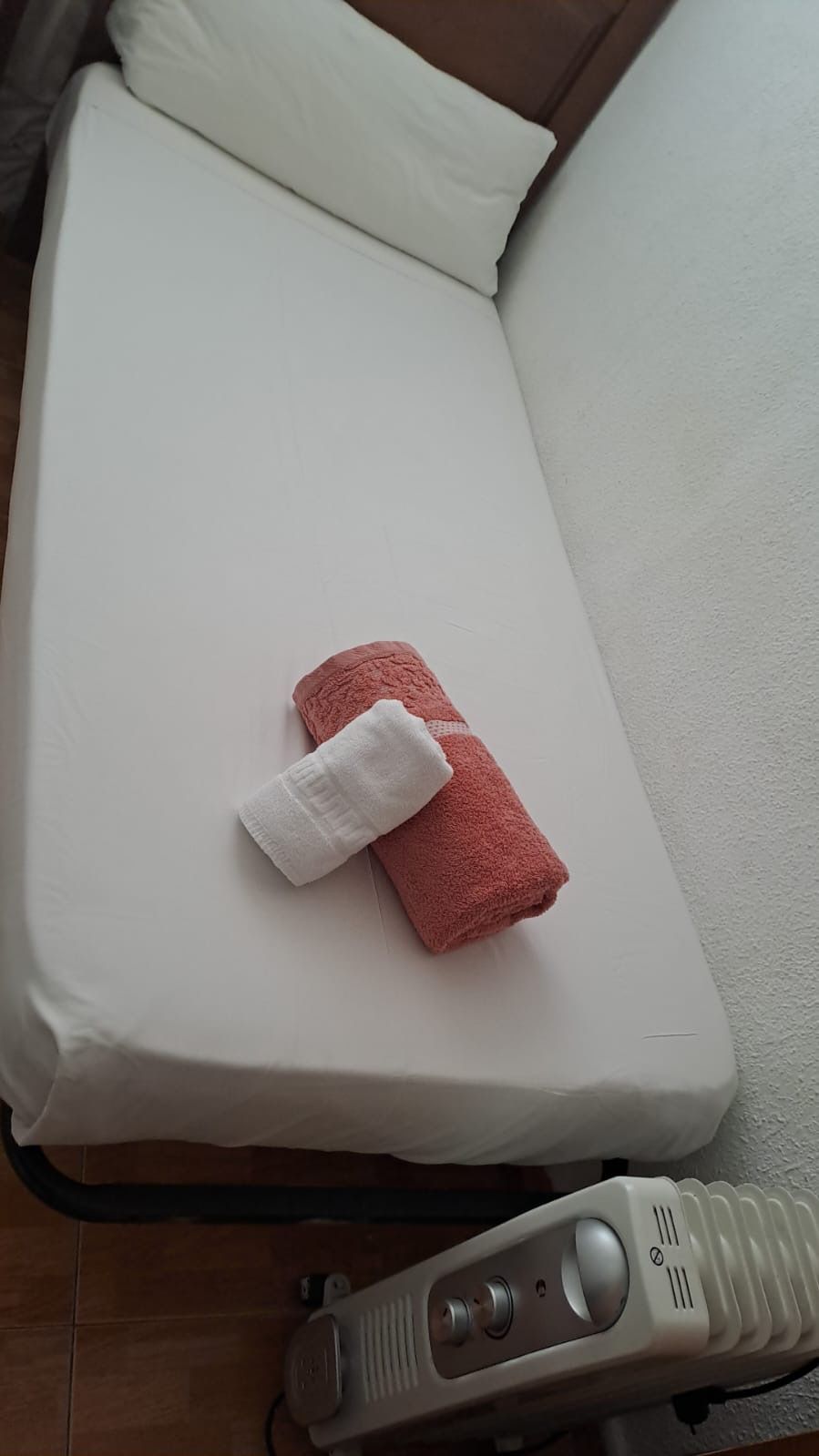 Cama con sábana blanca, almohada y toallas enrolladas, junto a una pared y calefactor.