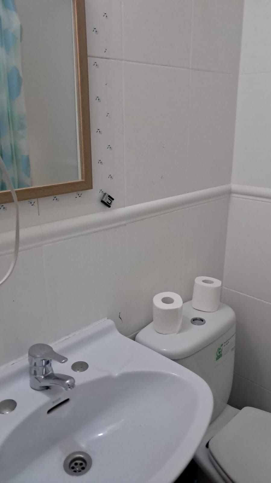 Baño con lavabo, inodoro, espejo y rollos de papel higiénico en la cisterna. Azulejos y paredes blancos.