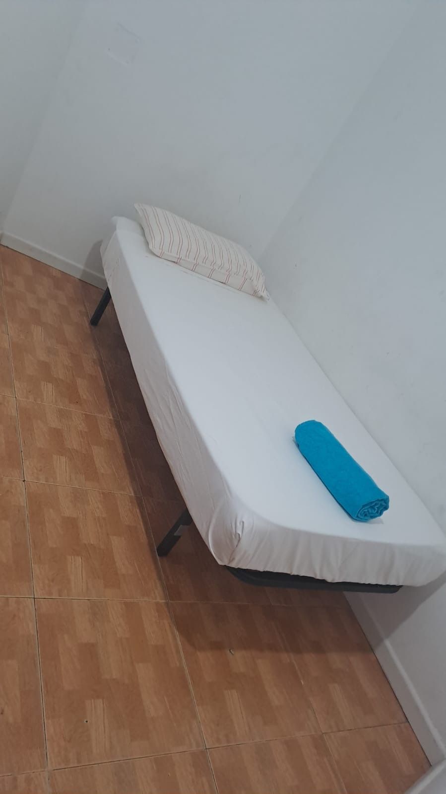 Una cama sencilla en una esquina con ropa de cama blanca, una toalla azul y una almohada estampada.