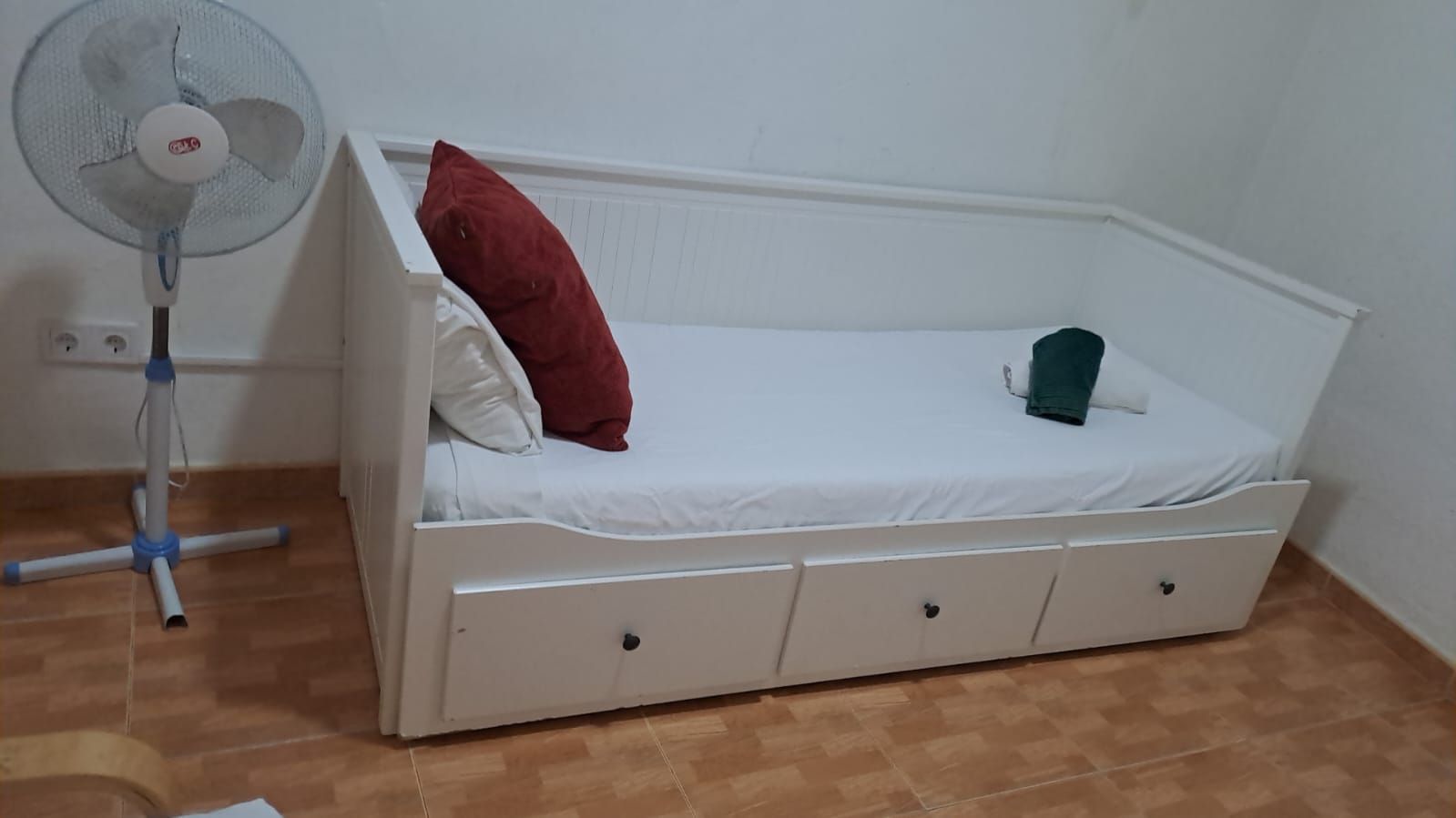 Sofá cama blanco con almohadas y cajones rojos y blancos, ventilador a la izquierda.