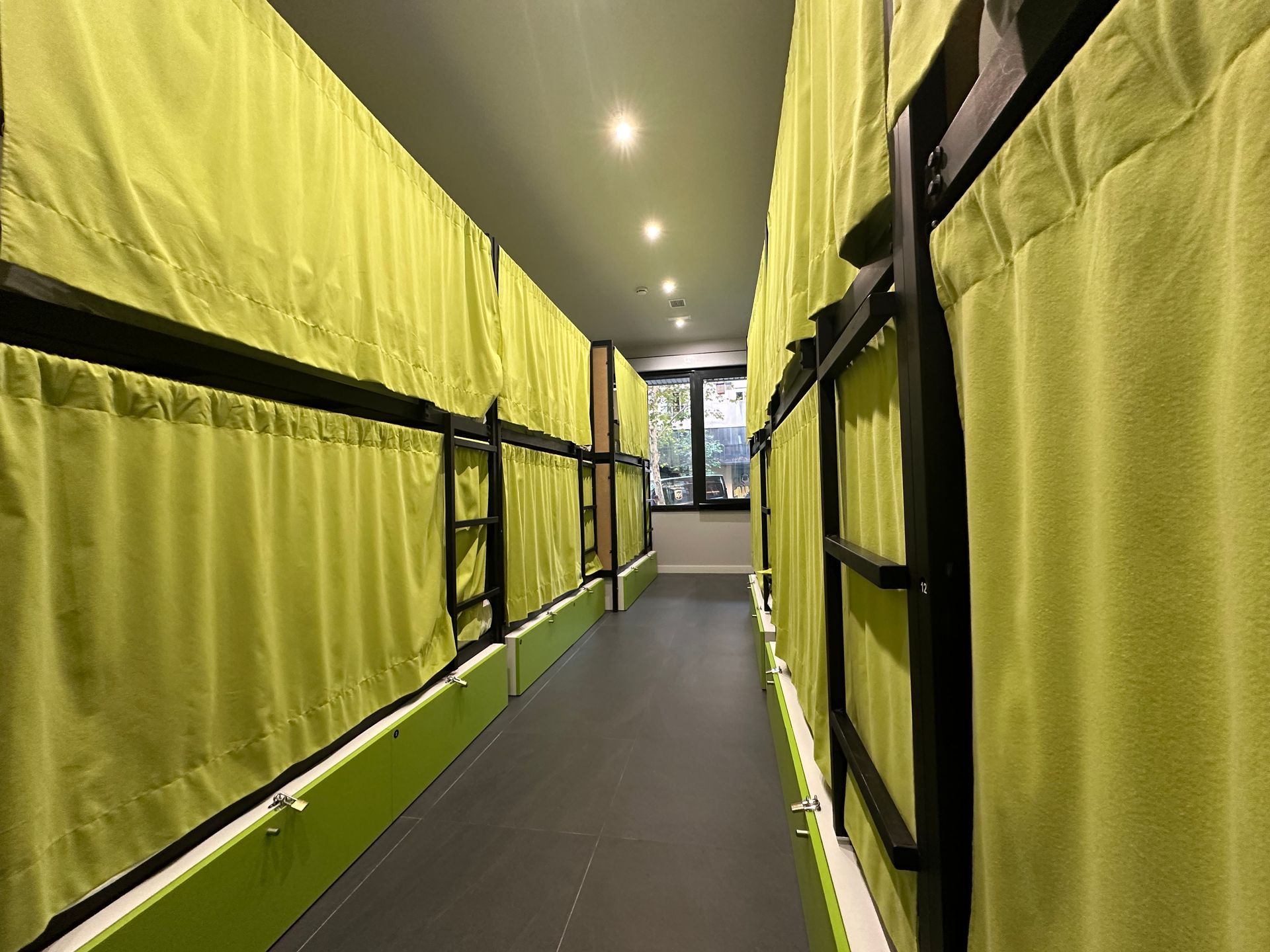 Un estrecho pasillo de albergue lleno de literas, cada una con una cortina verde.