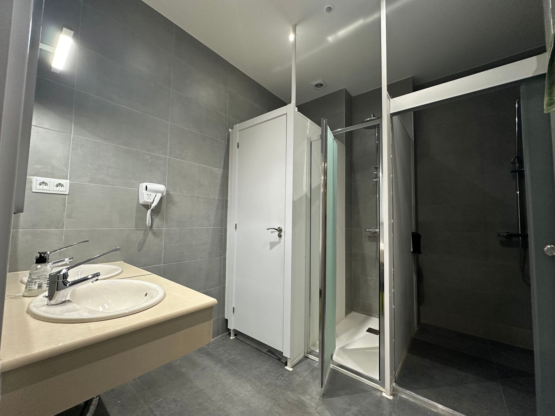 Baño con azulejos grises, lavabo blanco, ducha y puerta blanca.