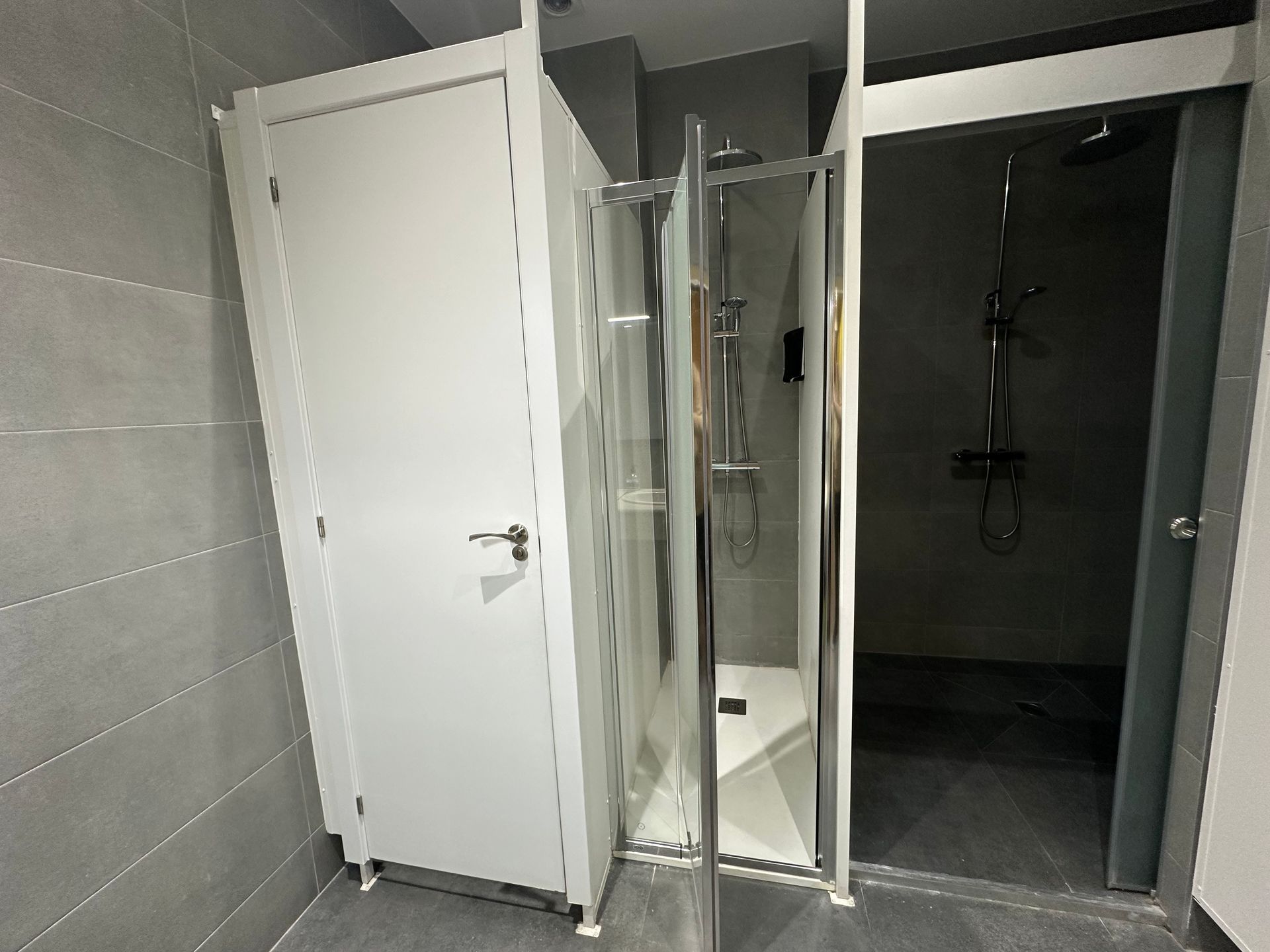 Baño con tres cabinas de color blanco, gris y cristal: inodoro, ducha y una ducha de color gris más oscuro.