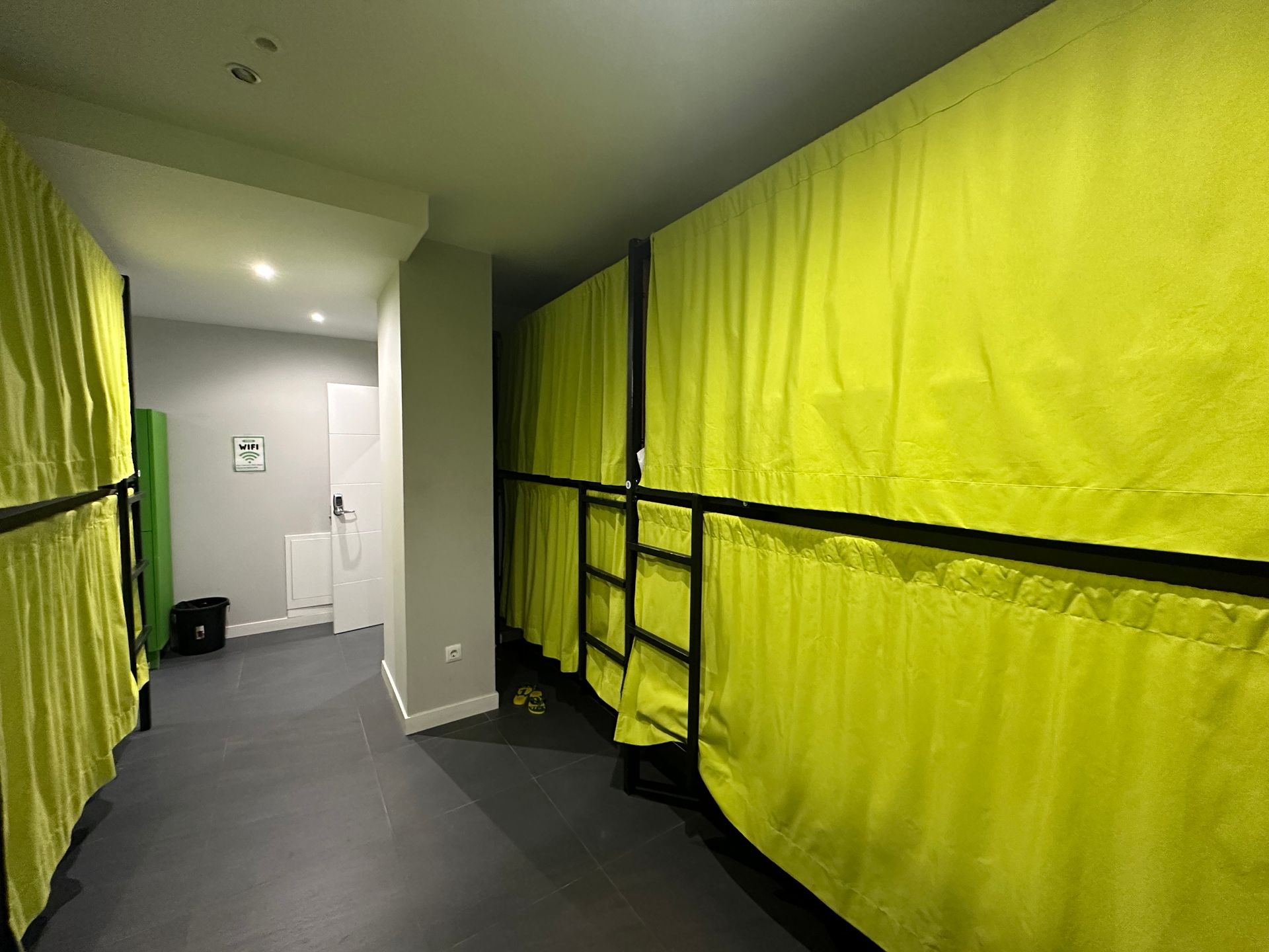 Una habitación de albergue con filas de literas, cada una con una cortina amarilla y un suelo gris oscuro.